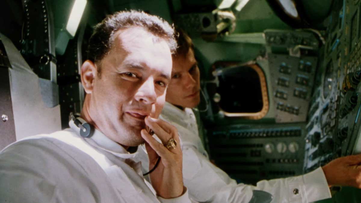 Still from the 1995 Apollo 13 - Two astronauts played by Tom Hanks and Kevin Bacon in a spacecraft interior. The foreground astronaut looks at the camera with a thoughtful expression, wearing a headset and white suit. The background astronaut is focused on the control panel. The setting appears to be a space mission.