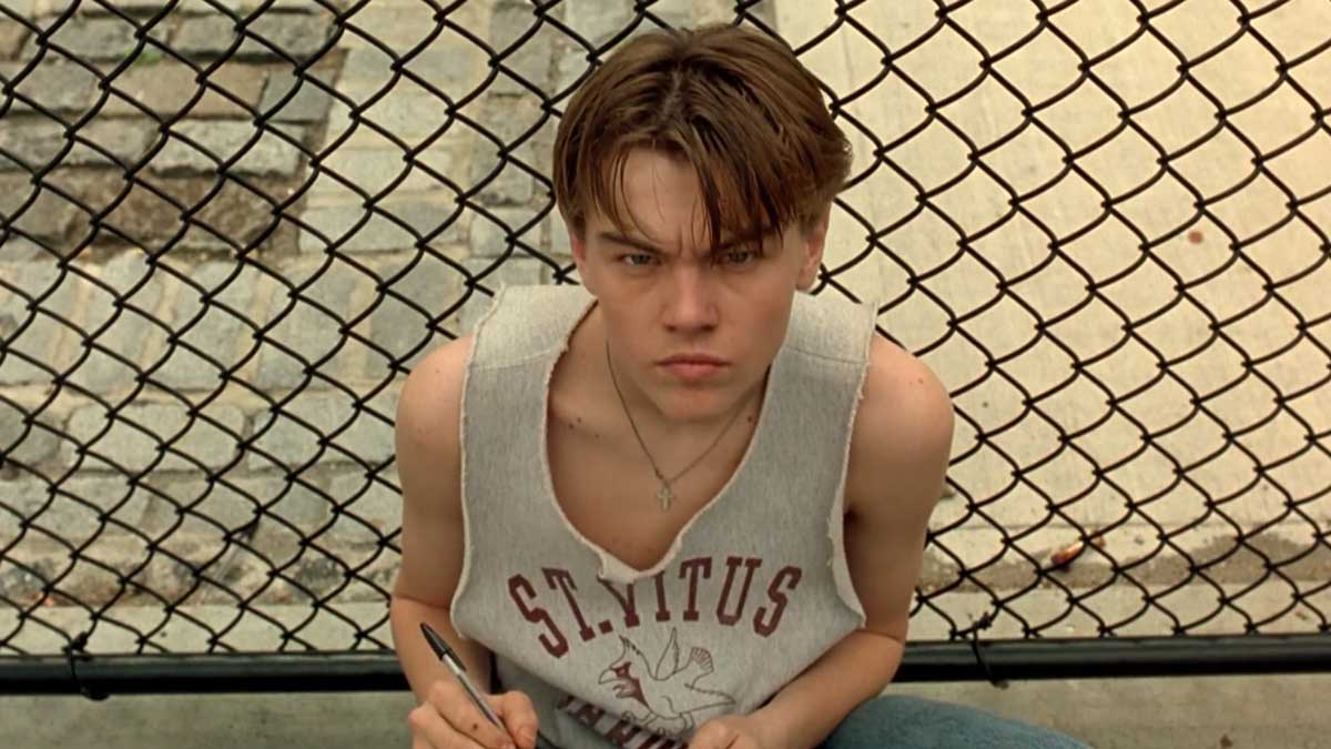 A young person with short hair sits in front of a chain-link fence. They are wearing a sleeveless shirt with St. Vitus printed on it and holding a pen and notebook, looking intently at the camera.