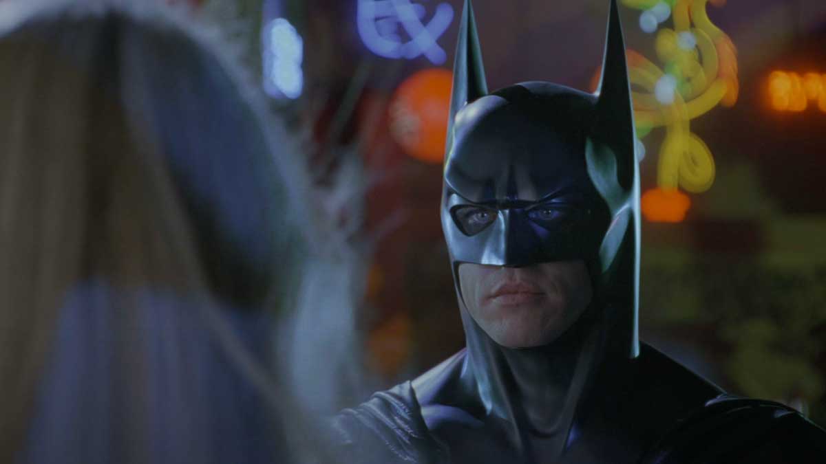 In a dimly lit setting with colorful neon lights, a person dressed as Batman, complete with a black mask and pointed ears, gazes at an individual with long hair. It's almost as if they're stepping out of one of those iconic 30 movies that turn 30 this year.