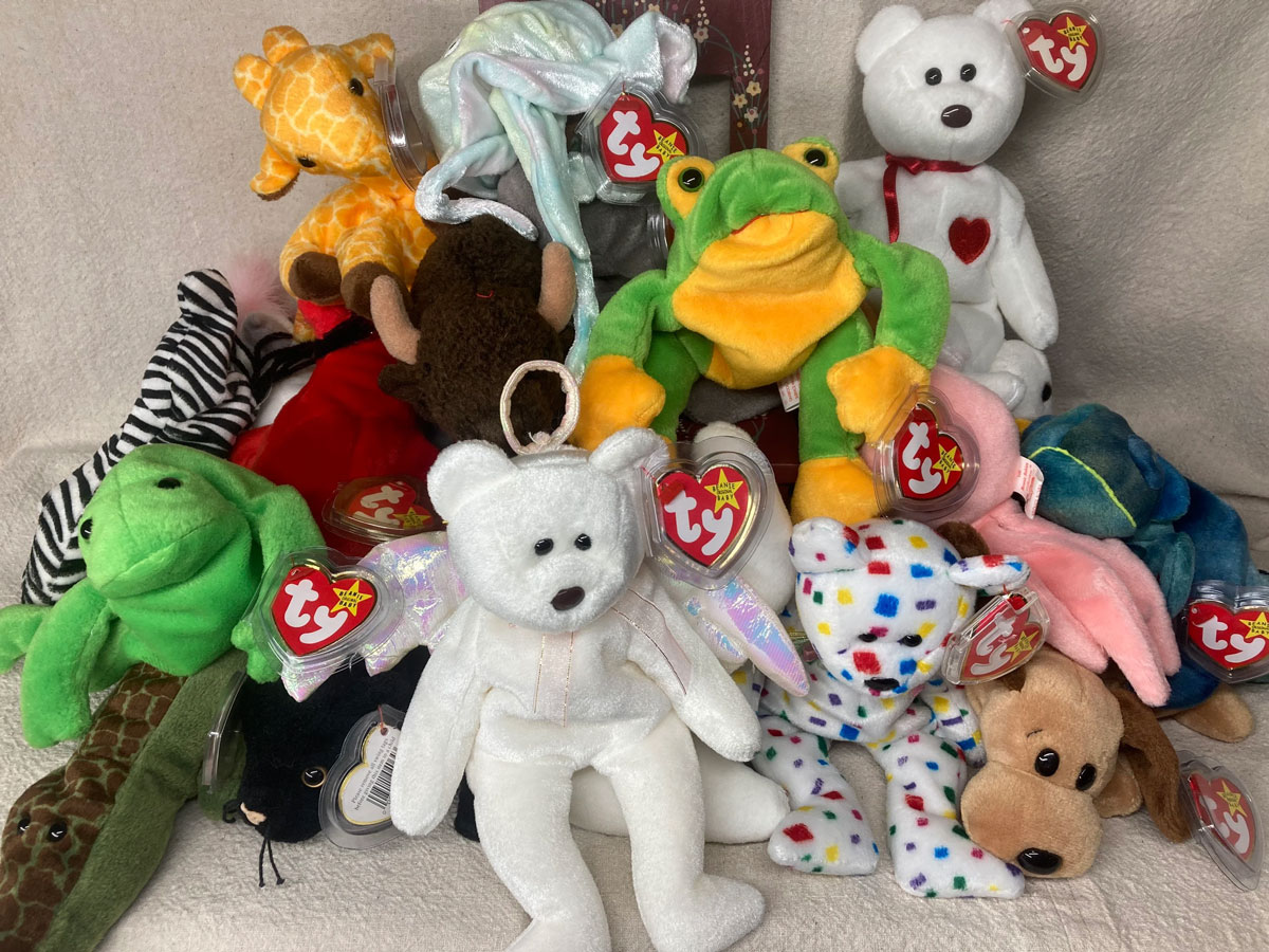 A collection of colorful stuffed animals, including bears, a giraffe, a frog, and a dog, with Ty tags attached, arranged on a soft beige surface.