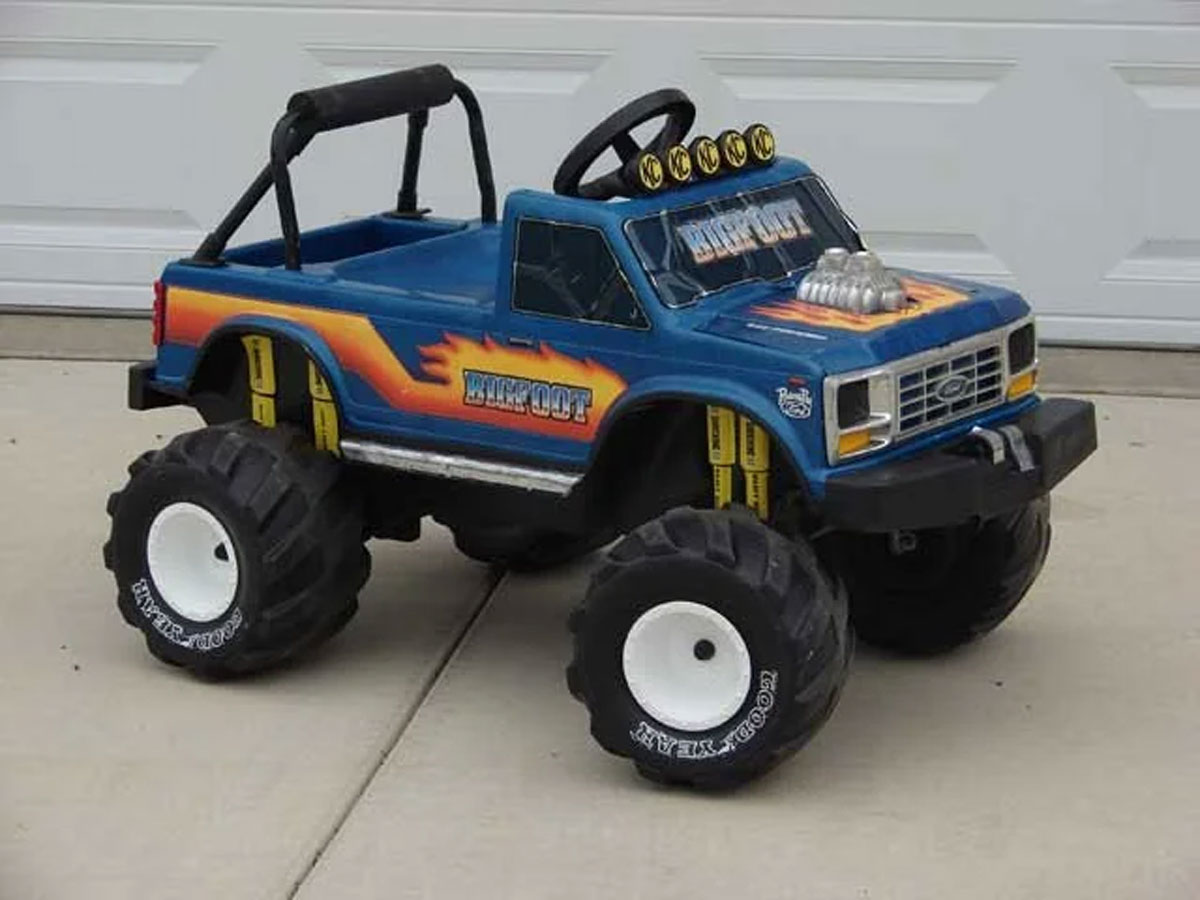 A blue toy monster truck with large black tires and white rims is parked on a concrete surface. It features yellow and orange flame decals, a black roll bar, and the word Bronco displayed on the front and side.