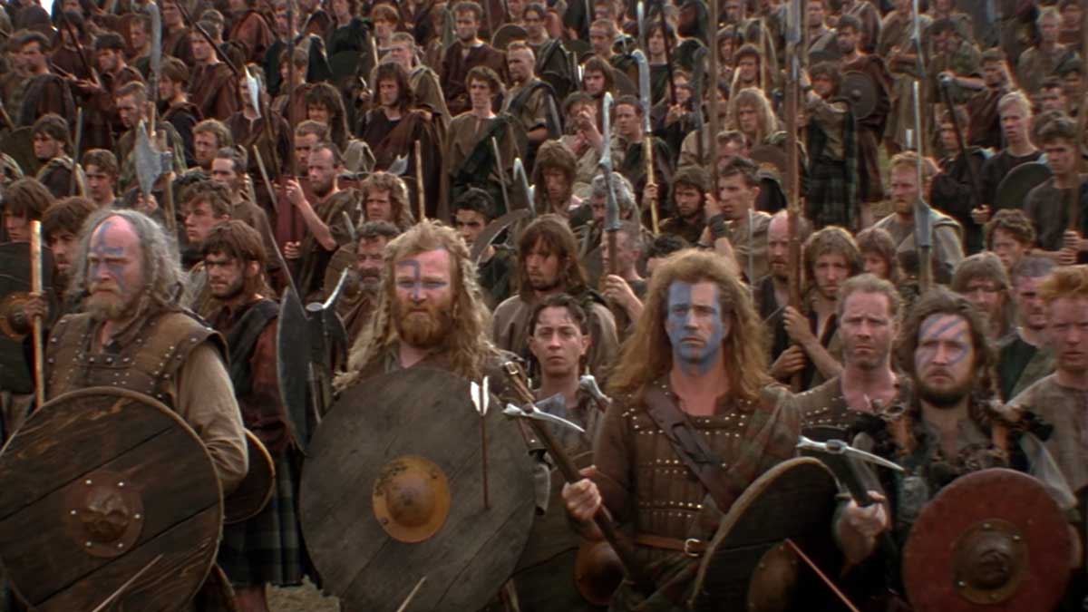 Still from the pre-battle scene in Braveheart. A large group of people in medieval-style clothing stand closely packed, holding shields and weapons. Mel Gibson's face is painted.