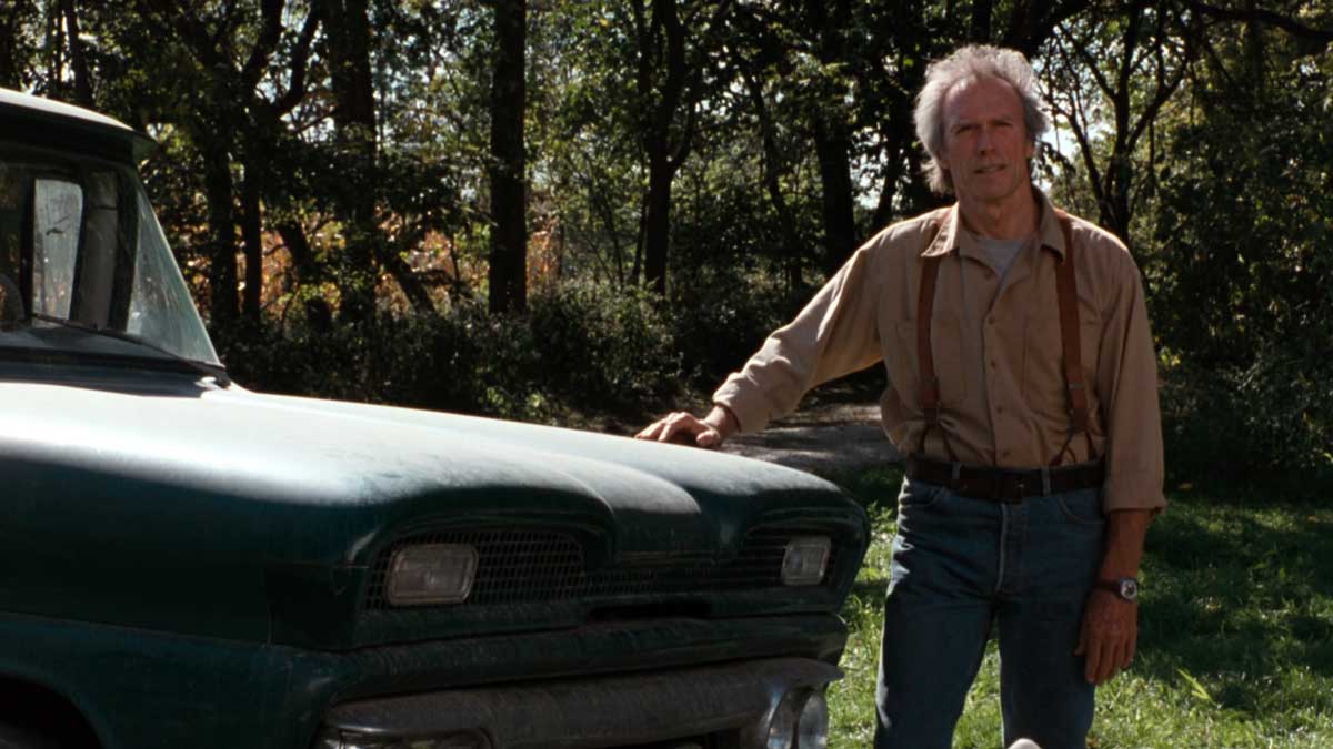 An older man stands outside next to the hood of a green classic truck. He is wearing a beige shirt and blue jeans with suspenders. The background is filled with lush green trees and grass under a canopy of sunlight.