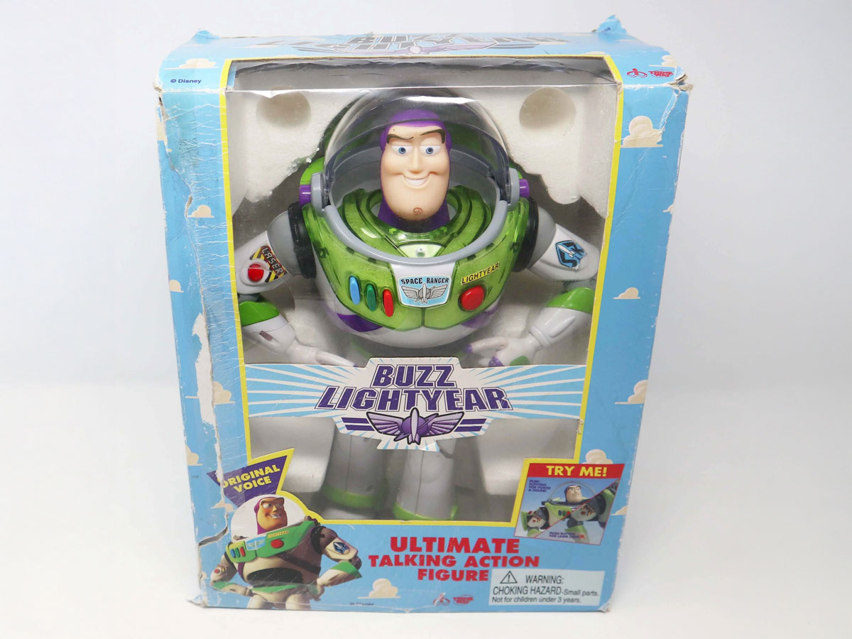 The image shows a boxed Buzz Lightyear talking action figure. The packaging is primarily blue with white clouds, featuring Buzz in a green and white spacesuit. It includes labels like Original Voice and Try Me!.