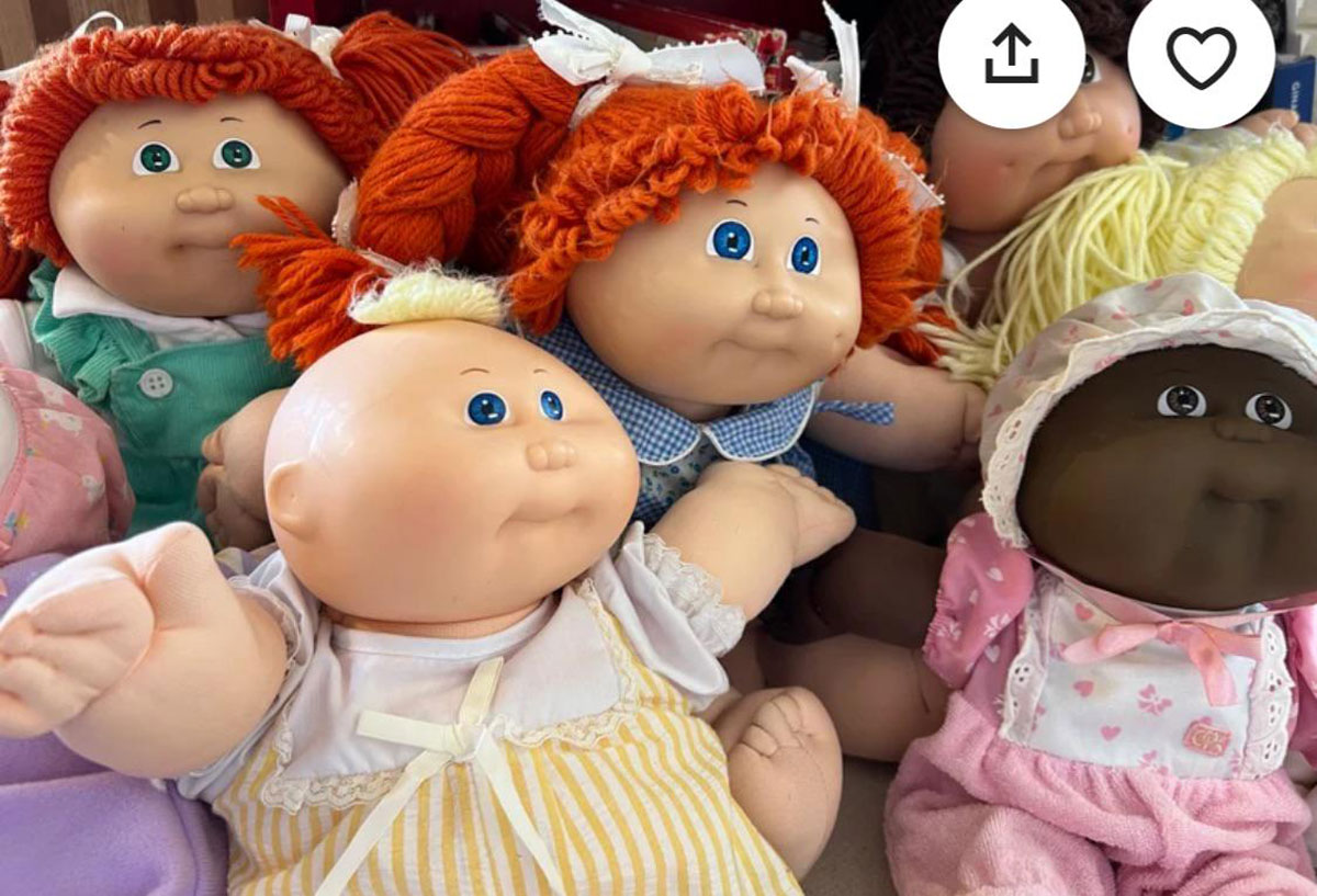 A collection of Cabbage Patch Kids dolls in various outfits and hairstyles, including red and blonde yarn hair, displayed closely together. The dolls have distinct round faces and big eyes, surrounded by various fabric textures and colors.