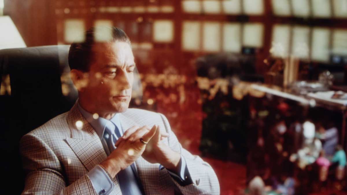 Casino 1995 - Robert De Niro in a suit sits contemplatively, with hands clasped and eyes seemingly focused on something in the distance. The scene has warm lighting, and reflections of a bustling casino are visible in the background glass.