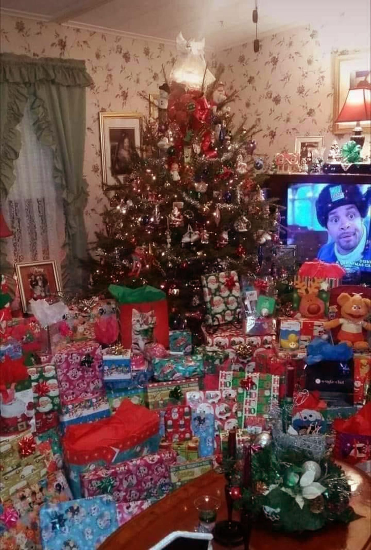 Huge pile of presents in front of a Christmas tree with Sinbad on the tv behind them