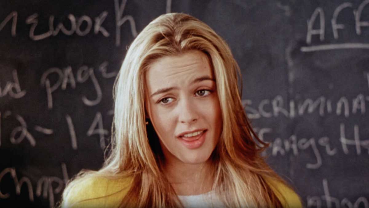 Alicia Silverstone in Clueless with long blonde hair stands in front of a chalkboard with handwriting on it. They are wearing a yellow jacket and a white top, looking slightly to the side with a relaxed expression.