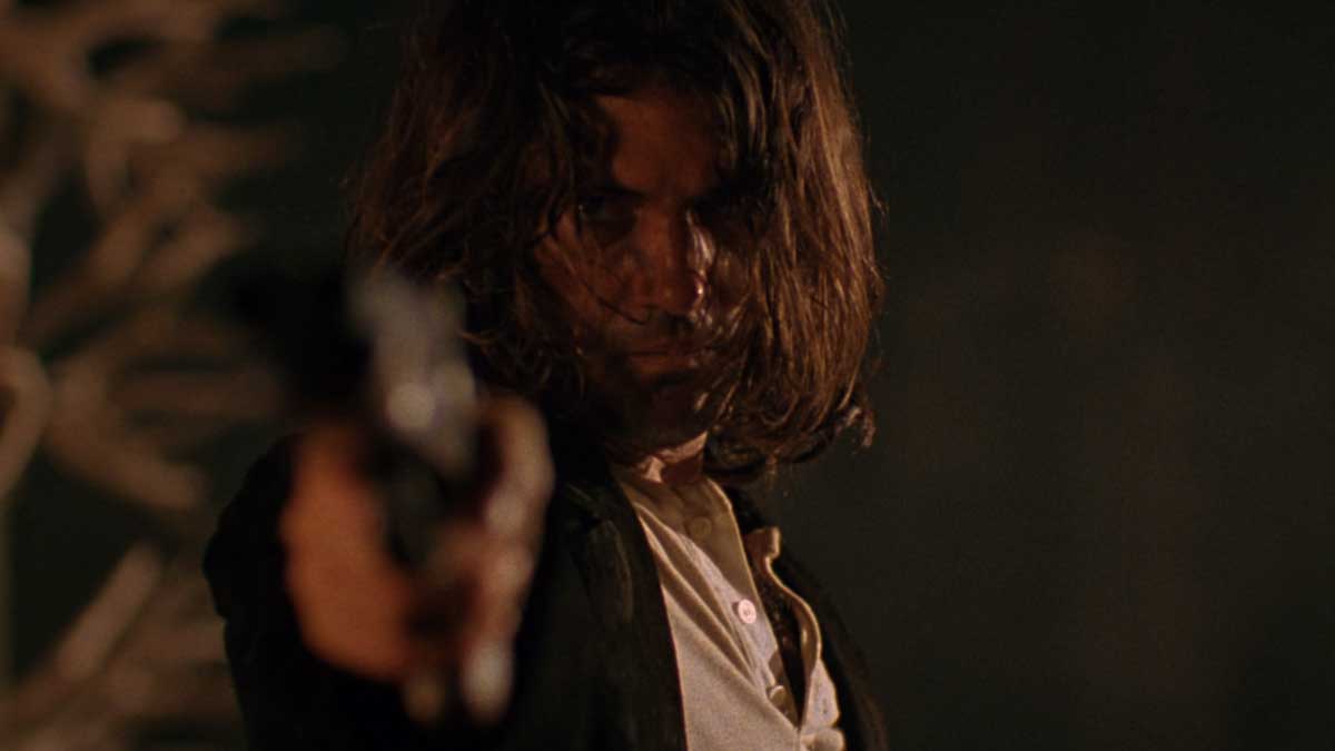 A person with long hair points a gun towards the camera in a dramatic and intense scene. The background is dark, enhancing the suspenseful mood.