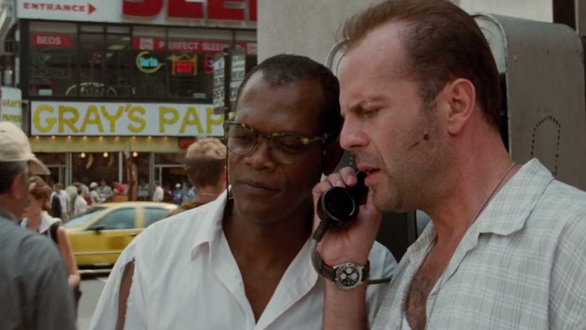 Two men stand close together on a city street. One man is speaking into a payphone, looking serious. They are both casually dressed. A yellow taxi is blurred in the background near a Grays Papaya sign.