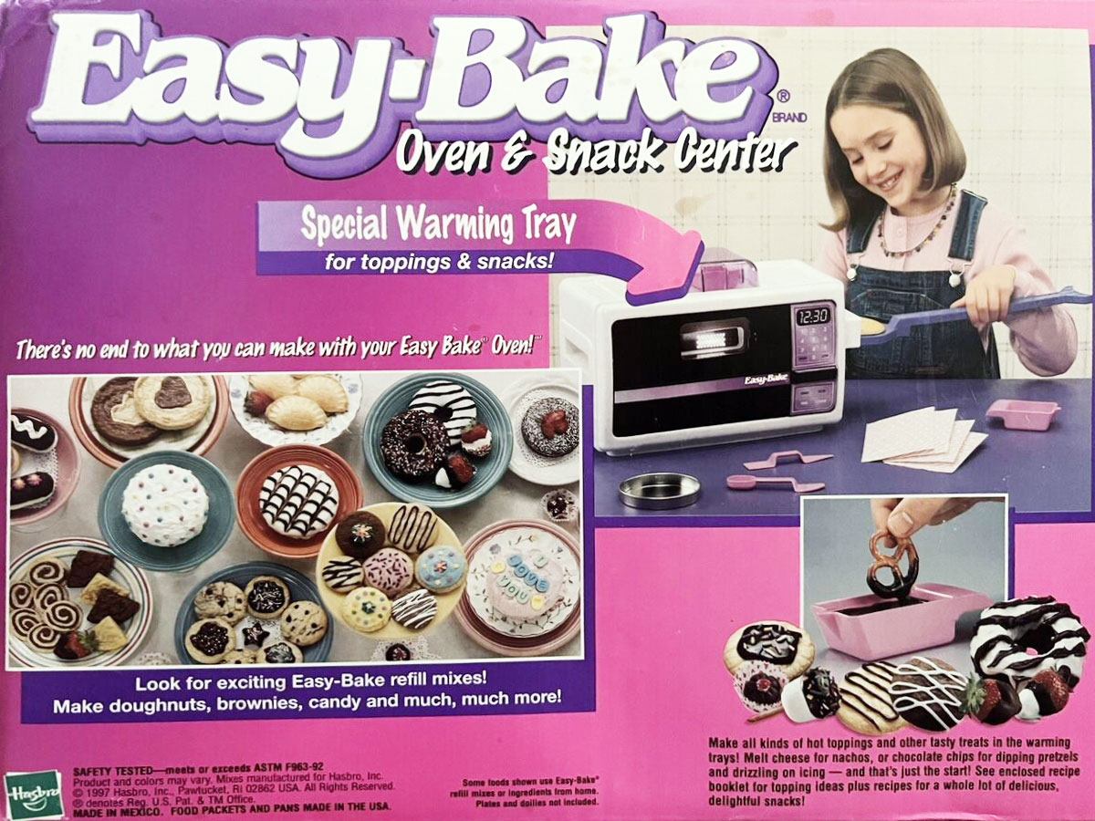 A girl uses the Easy-Bake Oven & Snack Center. The image shows various pastries like doughnuts and brownies on a special warming tray. The box highlights features like making doughnuts and includes refill mix options.