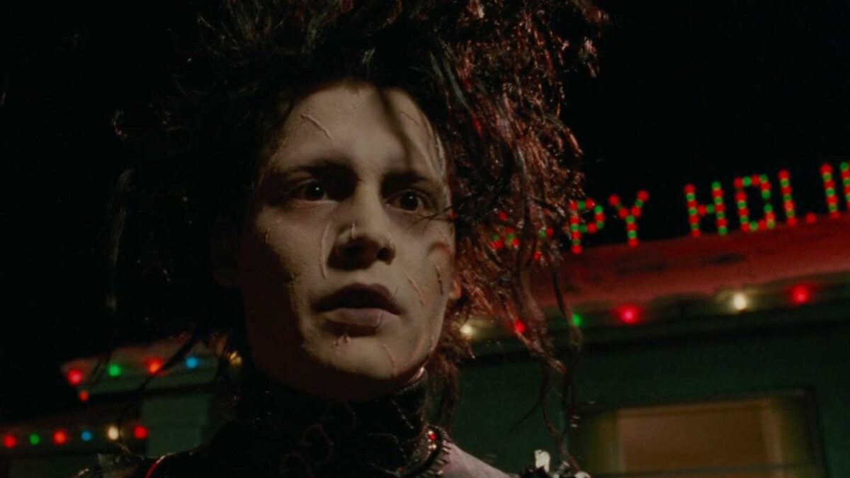 Cult Classic Christmas Movies - Edward Scissorhands - A person with wild black hair and pale skin, wearing dark clothing, stands in front of a building decorated with colorful holiday lights at night. The expression is somber, and the atmosphere is moody and dramatic.