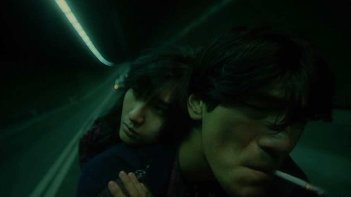 A person rides a motorcycle through a dimly lit tunnel. Another person is sitting behind them, holding on and looking over the rider’s shoulder. The rider has a cigarette in their mouth. The scene is enveloped in a greenish hue.