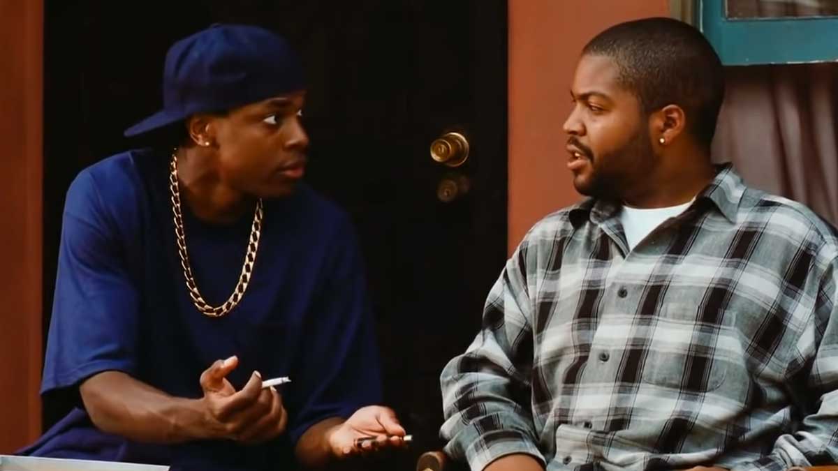 Cris Tucker and Ice Cube sitting and talking on a porch. The man on the left is wearing a blue shirt, hat, and gold chain, gesturing with his hands. The man on the right is in a plaid shirt, looking at him. A door with a brass doorknob is in the background.