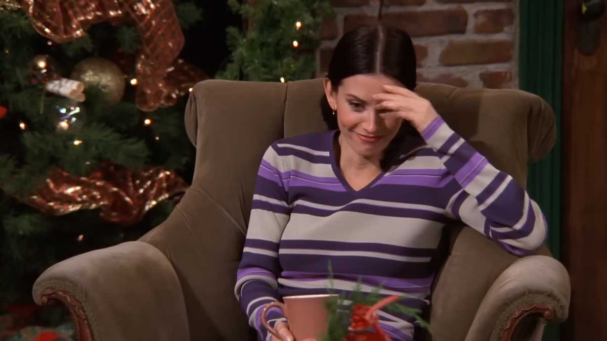 Christmas episodes of 90s tv shows - Friends - Monica sitting on a chair in Central Perk looking embarrassed. There's a Christmas tree behind her.