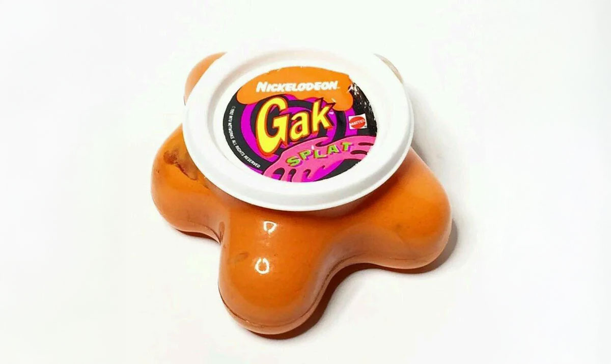 An orange splat-shaped container of Nickelodeon Gak with a white cap featuring colorful branding, lying on a white surface.