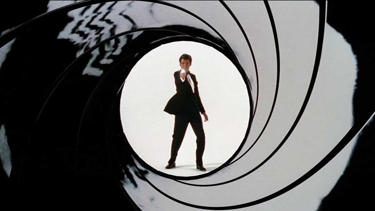 Pierce Brosnan as 007 in a suit points a gun toward the viewer, framed by a stylized spiral pattern resembling a gun barrel for the opening of Golden Eye. 