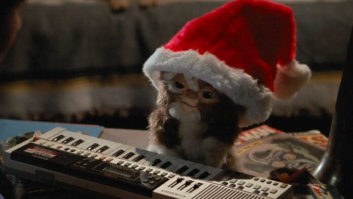 Cult Classic Christmas Movies - Mogwai wearing a Santa hat plays a small keyboard, with magazines and a chair in the background from the movie Gremlins.