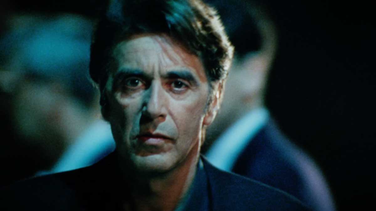 A man with an intense expression is captured in focus, looking directly at the camera. The background is blurred, highlighting his presence. The image has a moody ambiance with dim lighting, emphasizing dramatic features and emotions.