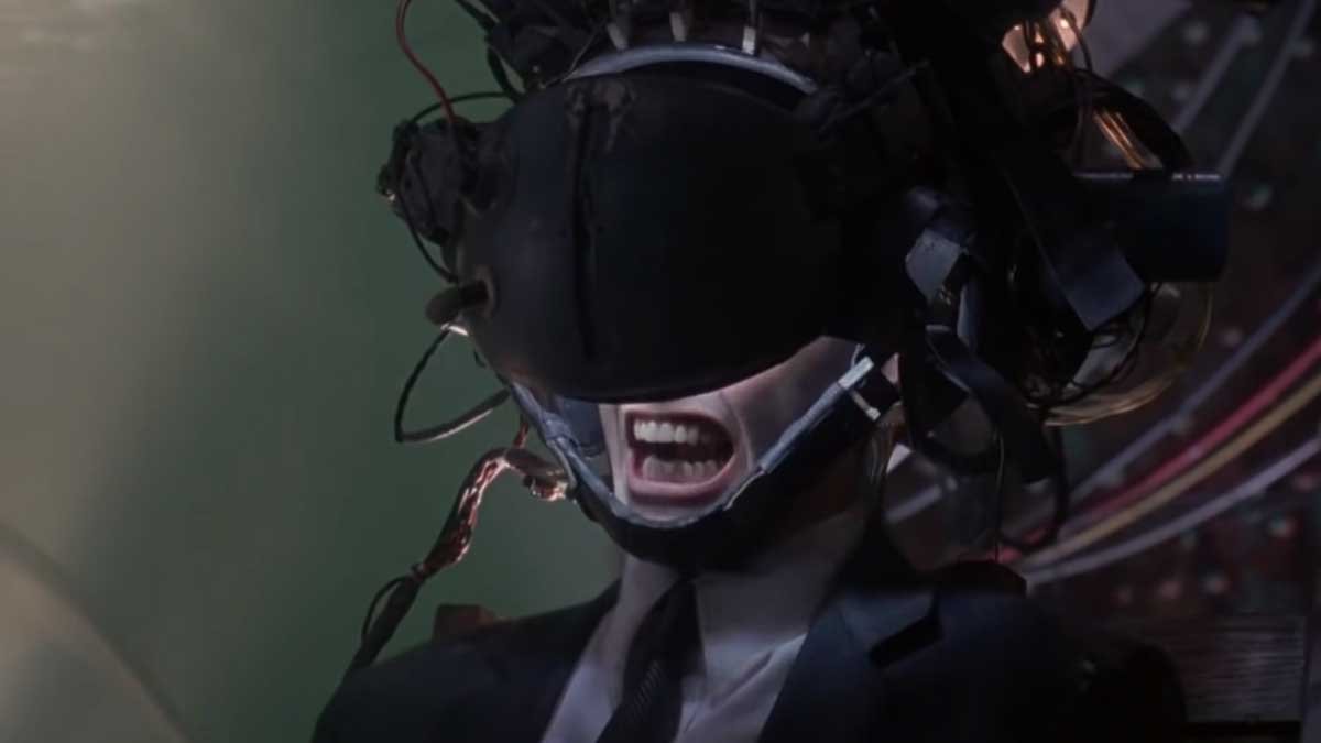 A person in a suit sits with a virtual reality headset and a complex tangle of wires and devices on their head. Their mouth is open wide in a scream or yell, conveying intense emotion or distress. The background is blurred and dark.