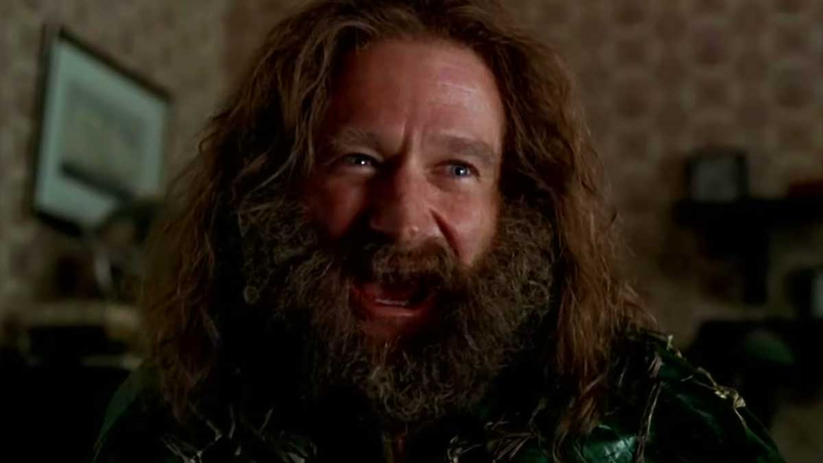 Robin Williams in Jumanji with long, unkempt hair and a thick beard is smiling widely. The background features a patterned wall and a framed picture. The scene has a warm, indoor lighting.