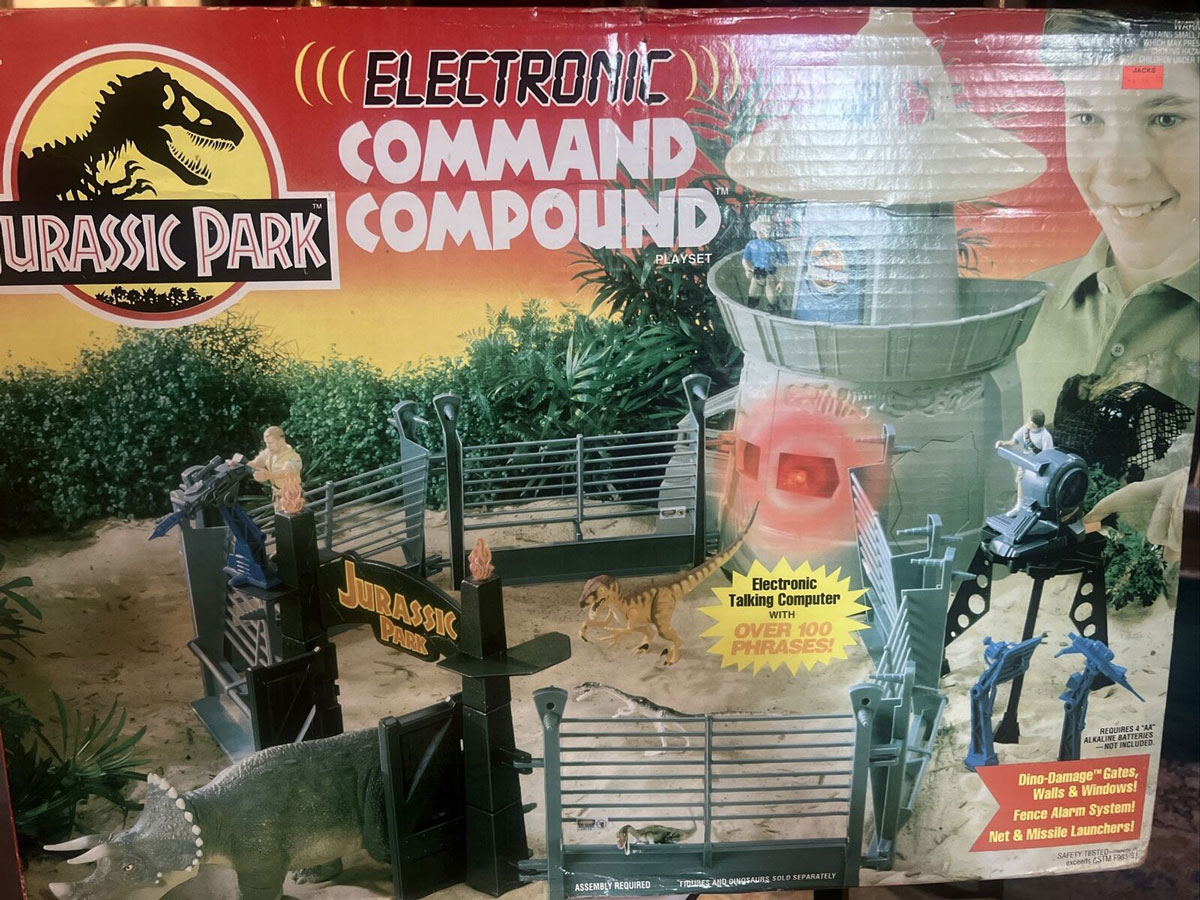 Box of a Jurassic Park Electronic Command Compound playset featuring dinosaur figures and a command tower. The box highlights over 100 phrases and includes accessories like an alarm and missile launcher. Background shows a dramatic dinosaur scene.