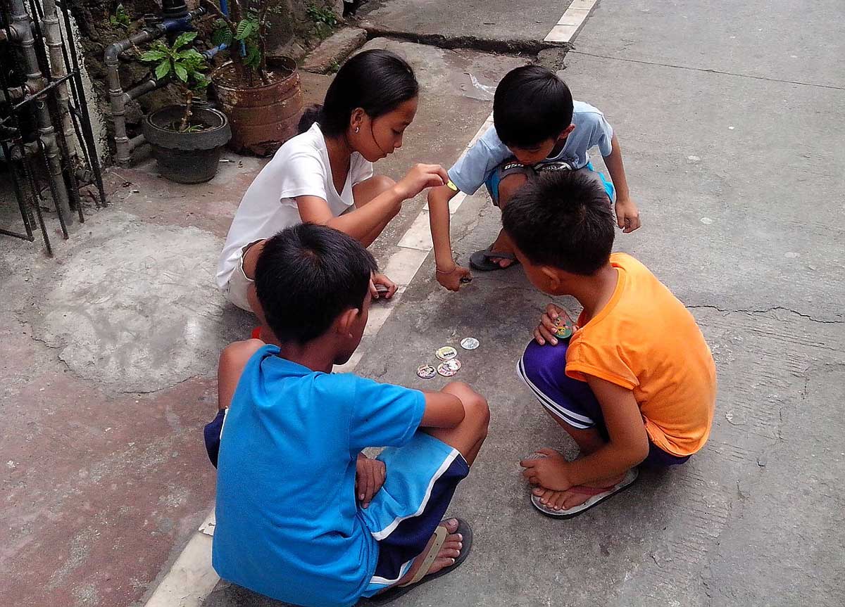 Four children are playing Pogs with circular paper pieces on a concrete street. They are intensely focused on the game, seated or crouched in a small circle. The surroundings include potted plants and a fence.