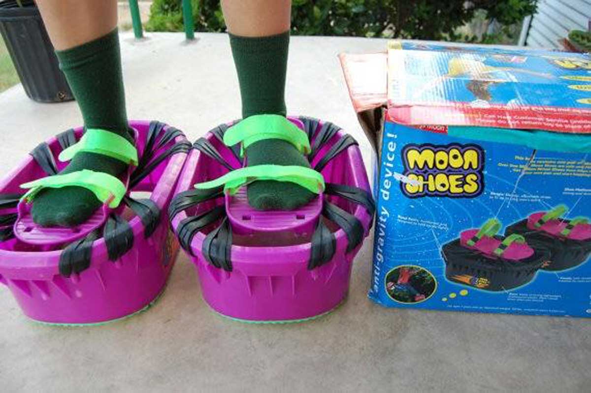 A person wearing green socks stands on purple Moon Shoes with green straps, next to the products colorful box on a concrete surface. The shoes are designed for bouncing, resembling mini trampolines for feet.