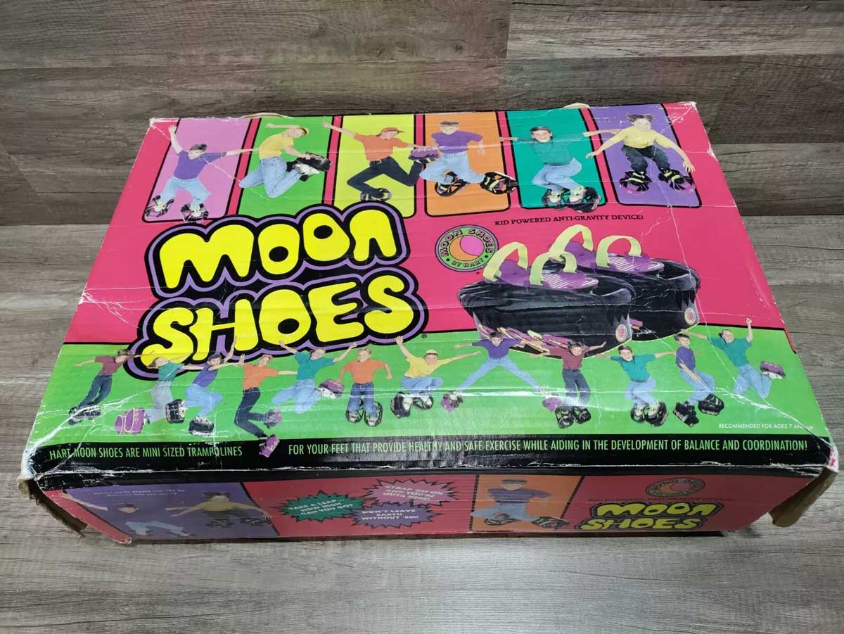 A colorful vintage box of Moon Shoes, featuring images of people jumping while wearing them. The box is red with green, yellow, and purple accents, and has the phrase Kid Powered Anti-Gravity Device printed on it.