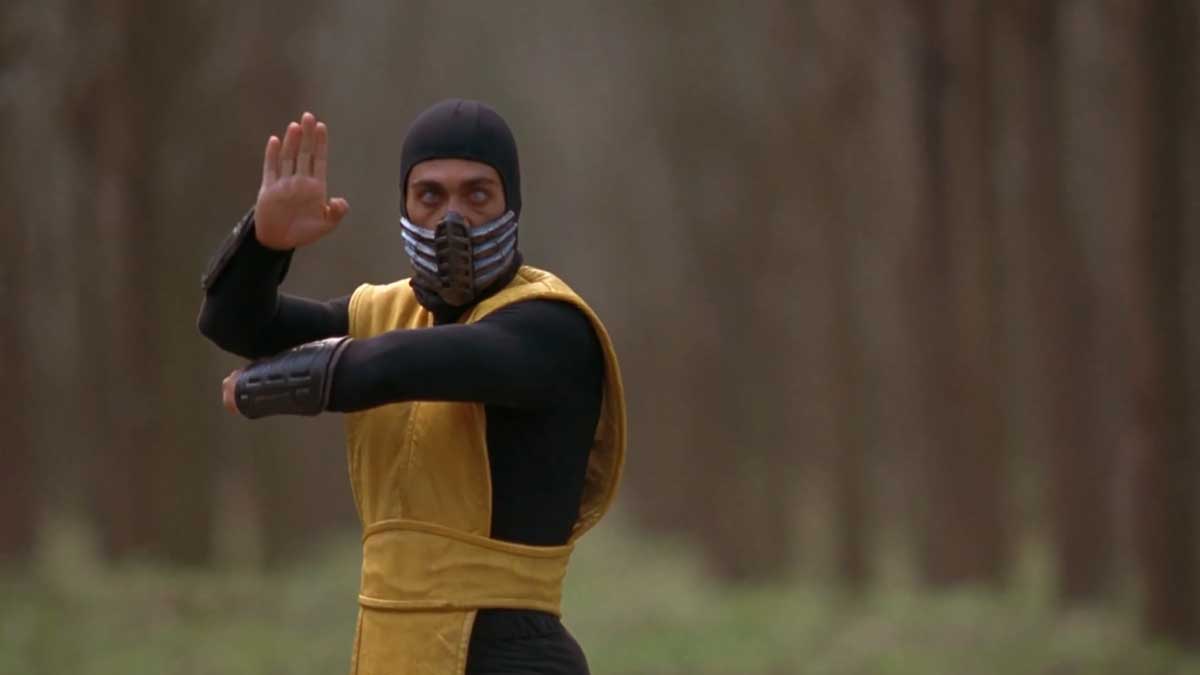 A masked martial artist wearing a yellow and black outfit poses in a forest setting. They have one arm raised defensively and the other extended forward. The background features blurred trees.