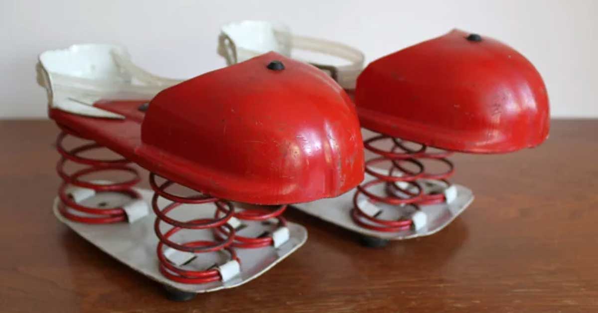 A pair of vintage red and white shoes with metal springs attached to the soles, designed for jumping, are placed on a wooden surface. The shoes have dome-shaped toe covers and a sturdy metal base.