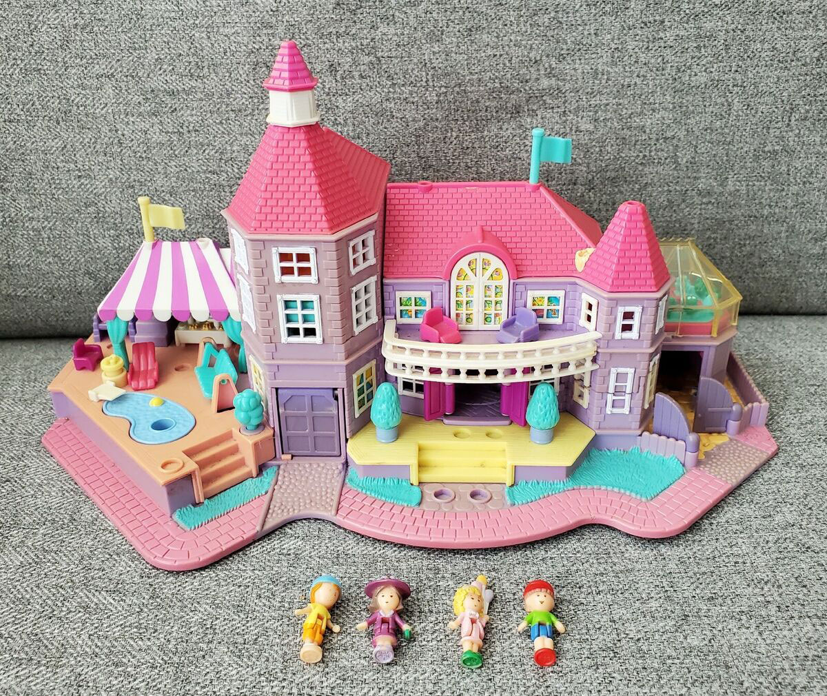 A colorful toy castle with pink and purple turrets, a pool, and a tent on a detailed play mat. Four small toy figures with varied outfits are lined up in front of the castle, all set on a gray textured background.