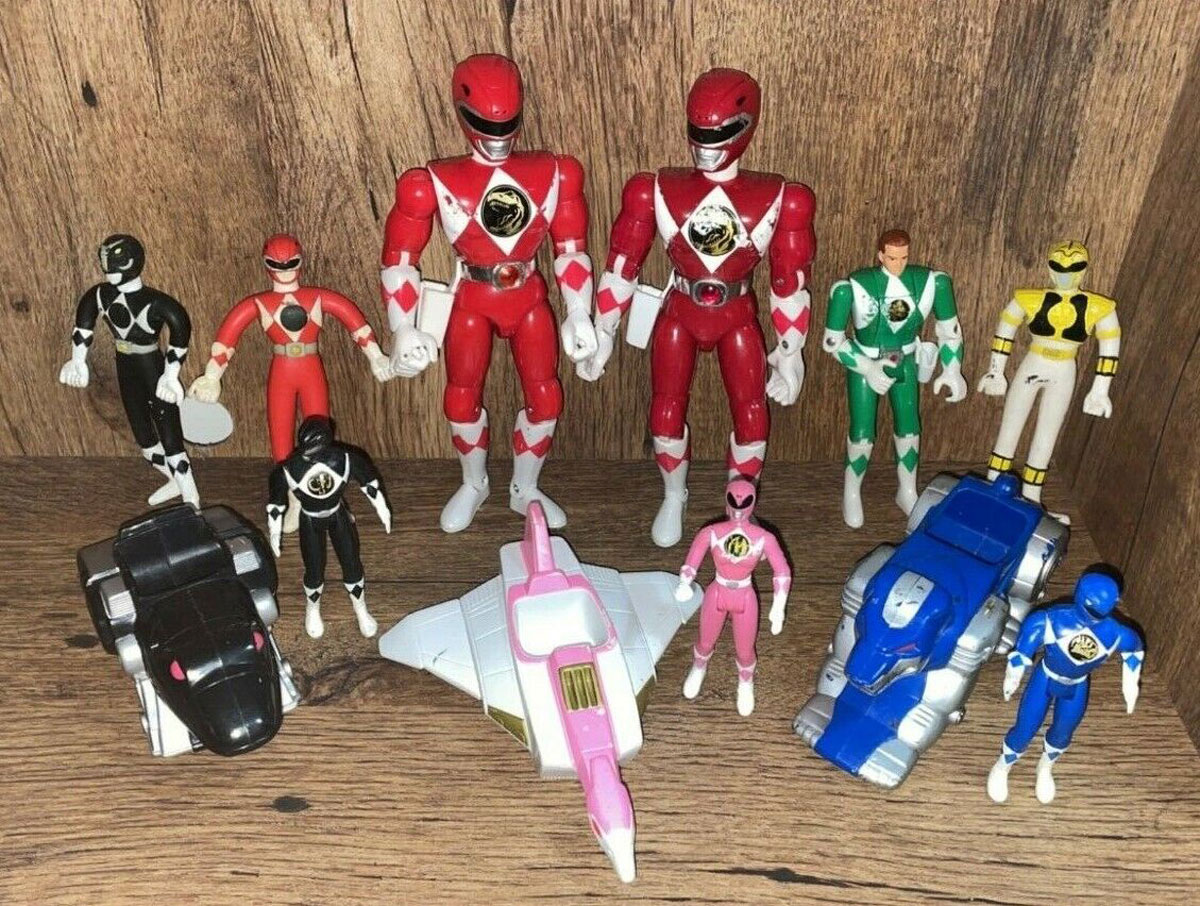 A collection of action figures featuring various Power Rangers in different colors, including red, black, pink, yellow, blue, and green, with accompanying toy vehicles, set against a wooden background.