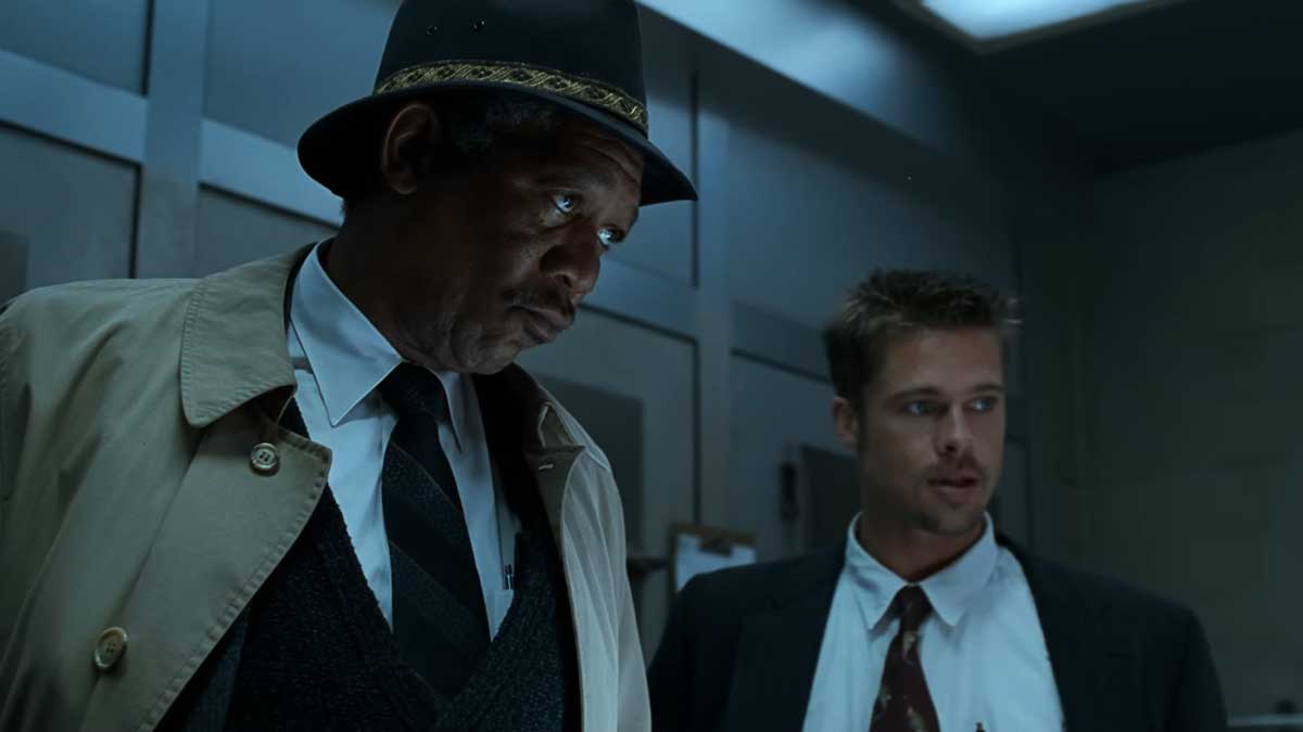 Morgan Freeman and Brad Pitt in the movie Se7en in suits are standing in a dimly lit room. The man on the left wears a hat and trench coat, looking serious. The man on the right, wearing a loose tie, appears attentive. The background is a plain, industrial setting.