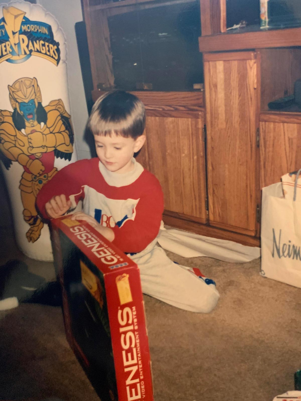 Kid unwrapping a Sega Gensis on Christmas morning in the 90s. Behind him is a Power Ranges toy.