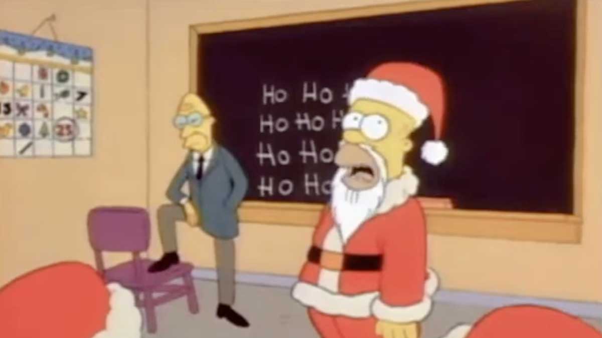 The Simpsons Christmas - Homer dressed up as Santa standing in front of a class of Santa's with the words Ho Ho Ho written on the chalkboard.