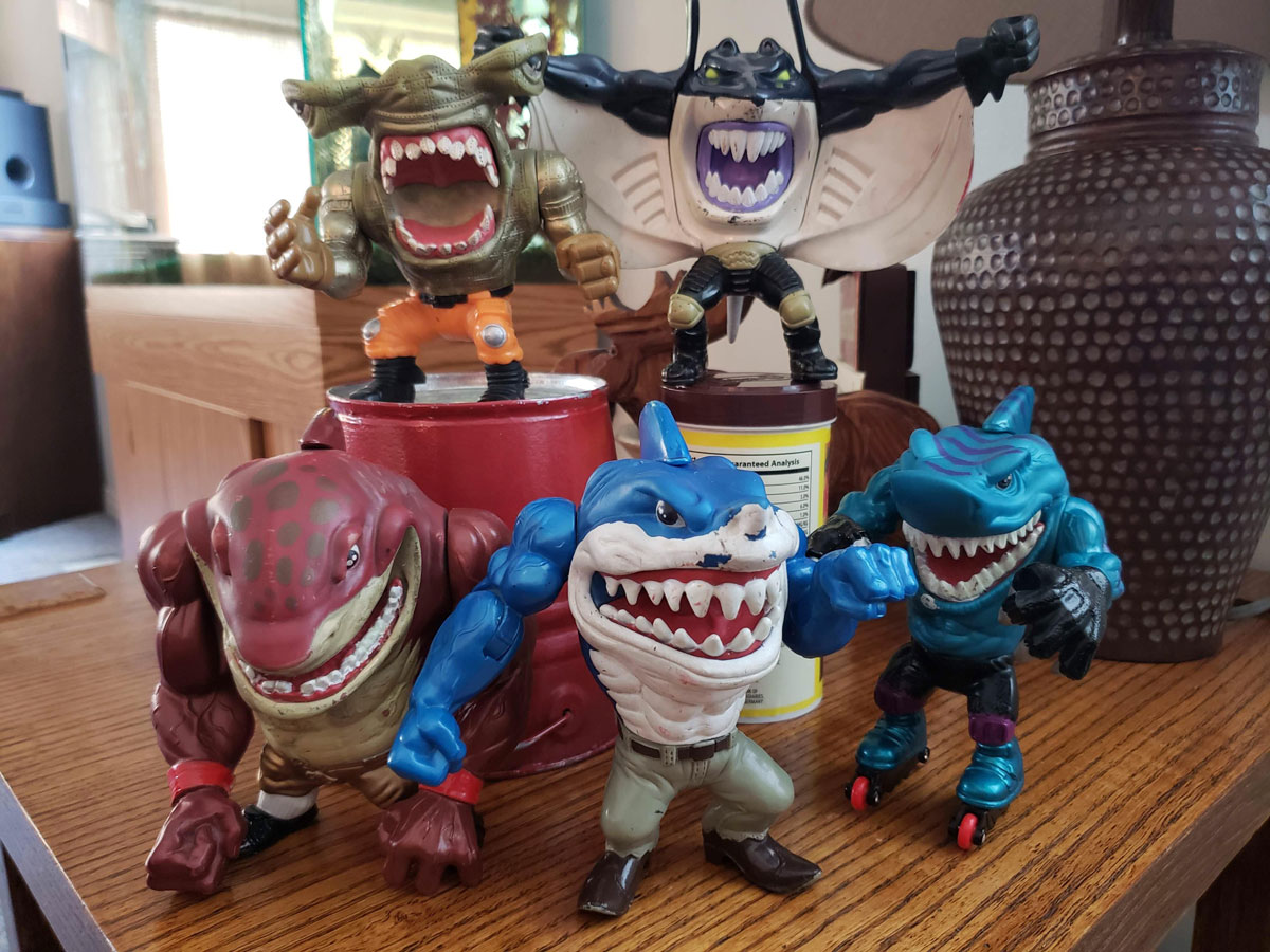 A group of five colorful shark-themed action figures with menacing expressions and exaggerated features are arranged on a wooden surface. Each figure has a unique pose and attire, including boxing gloves, roller skates, and fins.