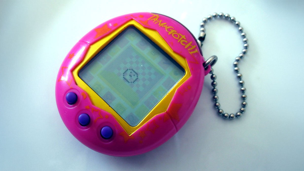 90s toys - A pink Tamagotchi device with purple buttons is attached to a silver beaded keychain. The screen displays a pixelated pet, and the word Tamagotchi is visible in bright yellow above the screen.