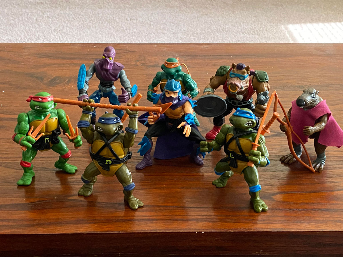 A collection of Teenage Mutant Ninja Turtles action figures is arranged on a wooden table. The figures include the four turtles, Splinter, Shredder, and other characters from the series, each in dynamic poses and equipped with various weapons.