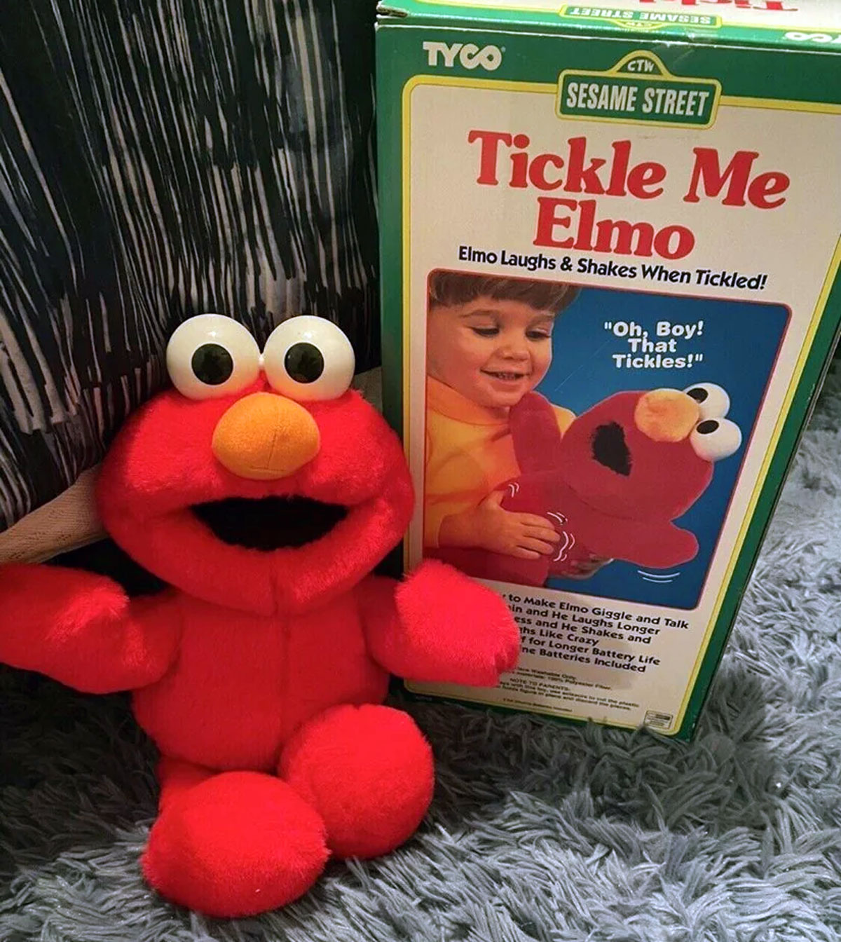 A Tickle Me Elmo plush toy is seated on a gray carpet next to its packaging box. The box features a child demonstrating how Elmo shakes when tickled. Elmo is bright red with big eyes and an orange nose.