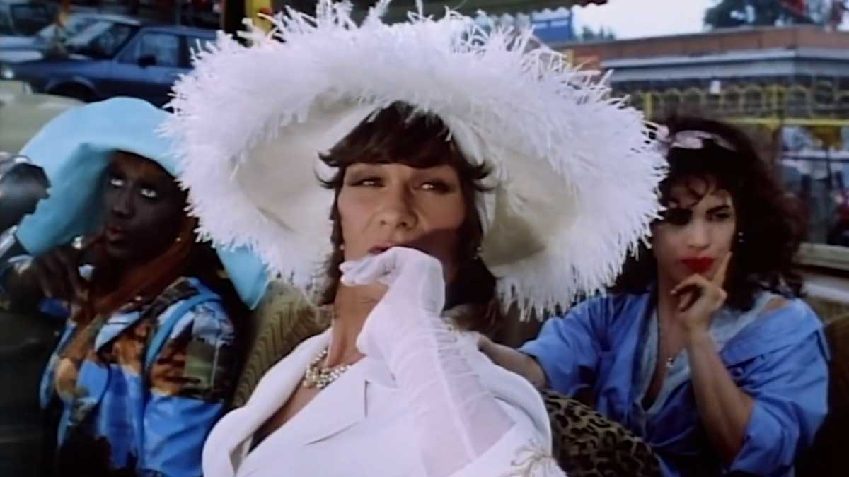 Three people sitting in a car. The person in the middle wears a large white hat and white gloves, appearing contemplative. The person on the left wears a blue hat, and the person on the right has curly hair and touches their mouth pensively.