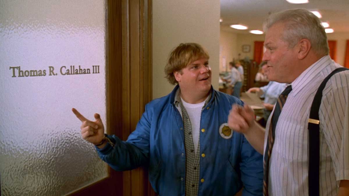 Two men stand in front of a door labeled Thomas R. Callahan III. The man on the left, wearing a blue jacket, points at the name. The man on the right, in a white shirt with suspenders, looks at the other man. An office setting is visible in the background.