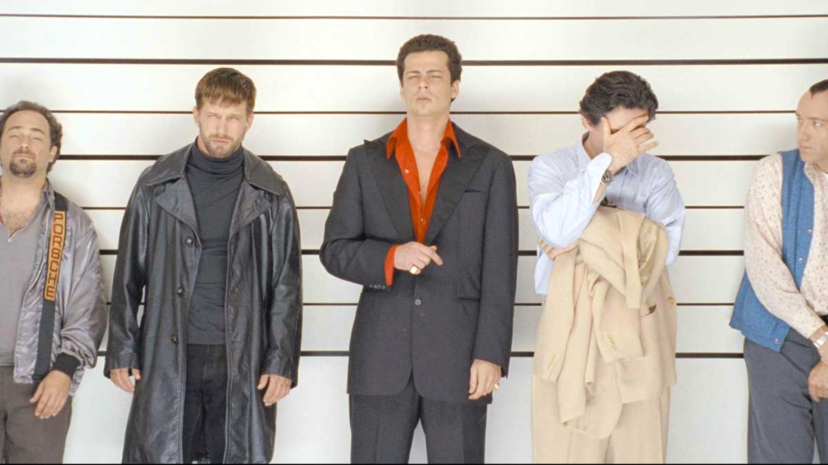 Five men stand against a lineup wall in The Usual Suspects. Each shows a different expression and attire, ranging from casual to formal. One man wears a long coat, another a suit, while another covers his face with his hand.