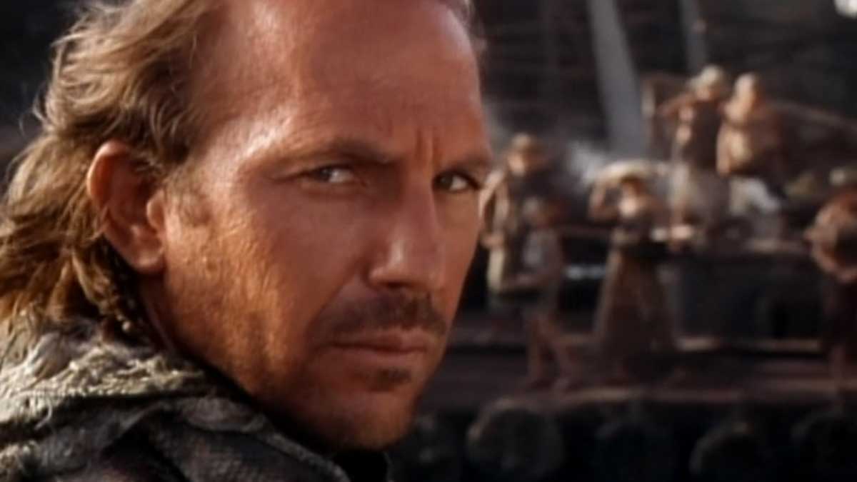 Close-up of a man with long hair and a serious expression, standing on a boat. The background shows blurred figures and nautical equipment, suggesting a maritime setting.