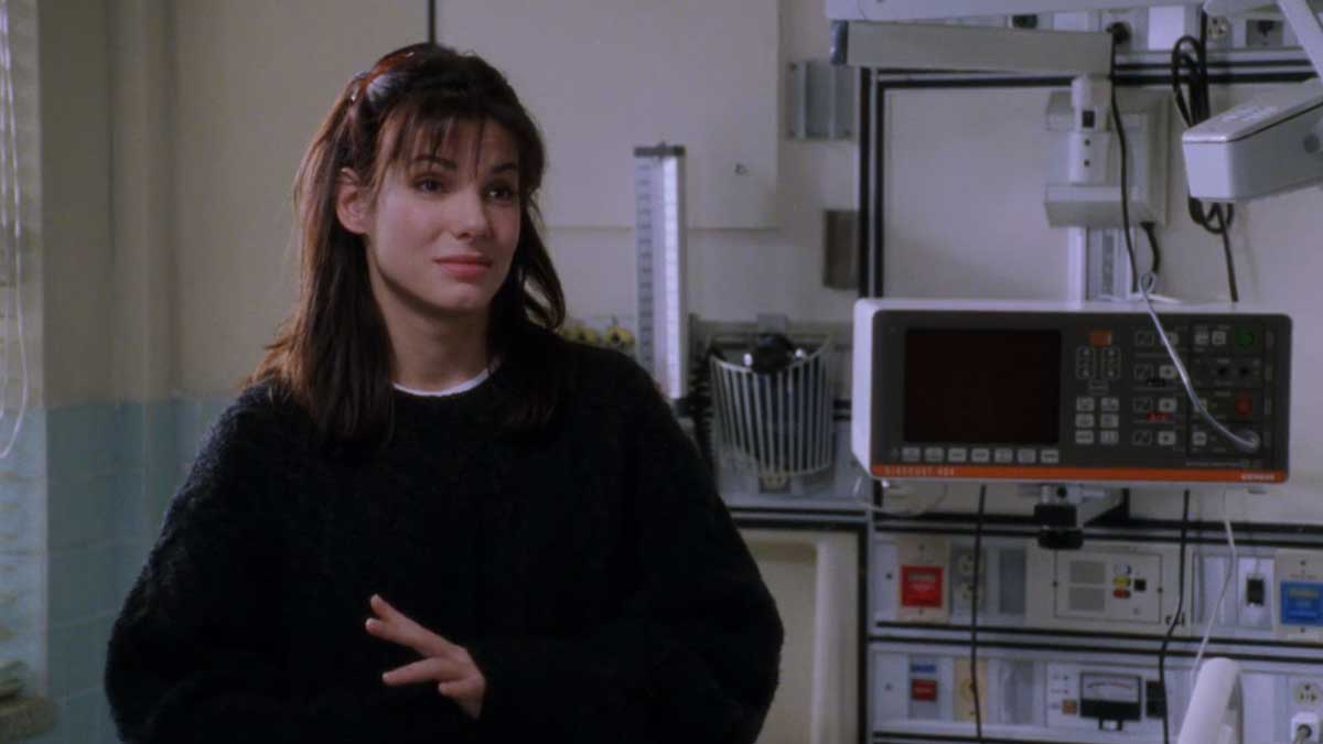 A woman with dark hair stands in a medical setting, wearing a black sweater. She is near medical equipment on the wall. The room is lit by natural light from a window.