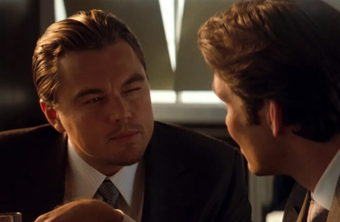 Leonardo DiCaprio man squints slightly while looking at another man during a conversation. Both are wearing suits. The background is dimly lit, suggesting an indoor setting. Still from Inception.