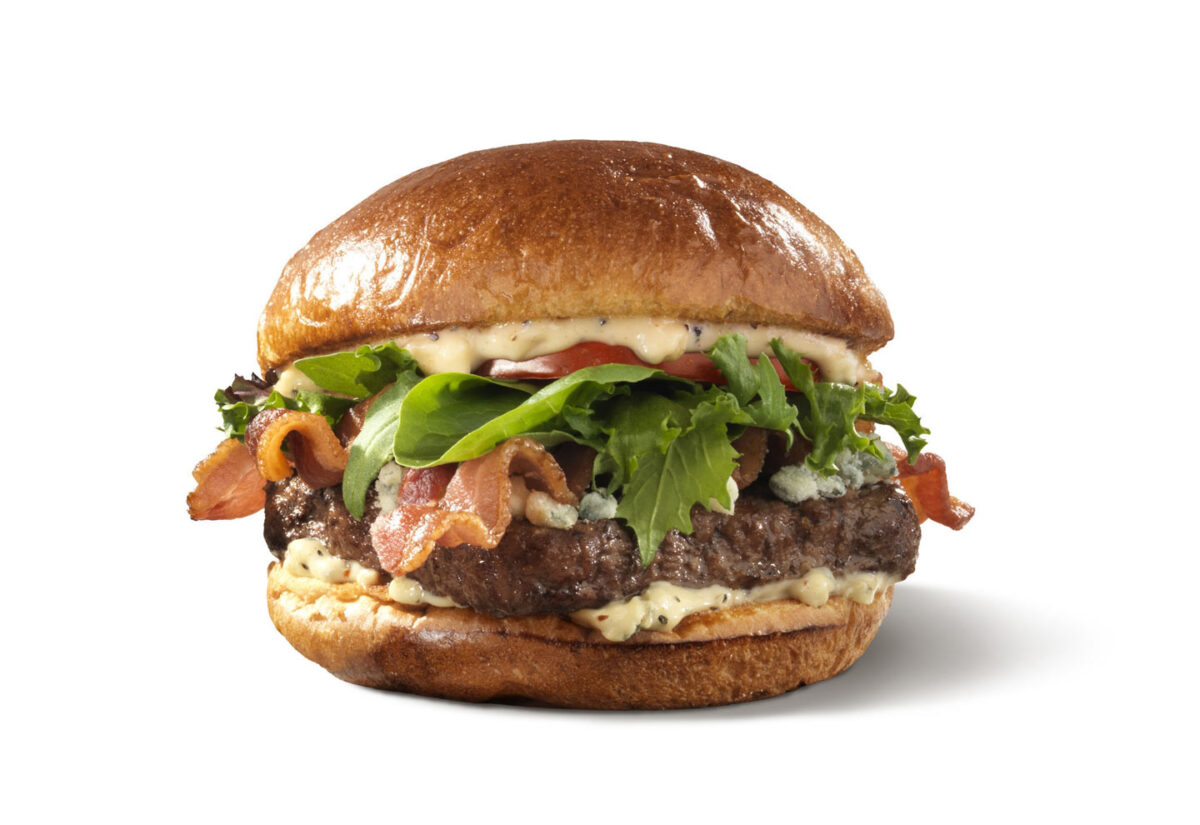 A hamburger with a glossy bun, topped with leafy greens, slices of bacon, and a thick beef patty. The burger is garnished with mayonnaise-based sauce and crumbled blue cheese, on a plain white background.