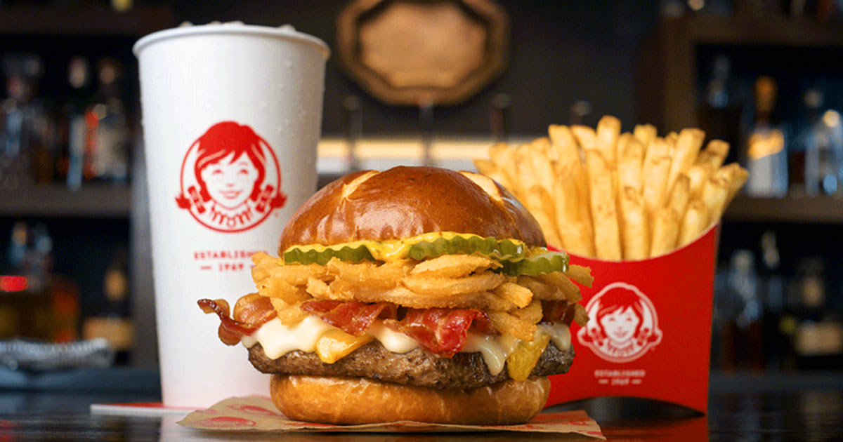 A burger with bacon, cheese, crispy onions, and pickles on a bun is in the center. French fries are served in a red container on the right, and a large drink in a white cup with a red logo is on the left.
