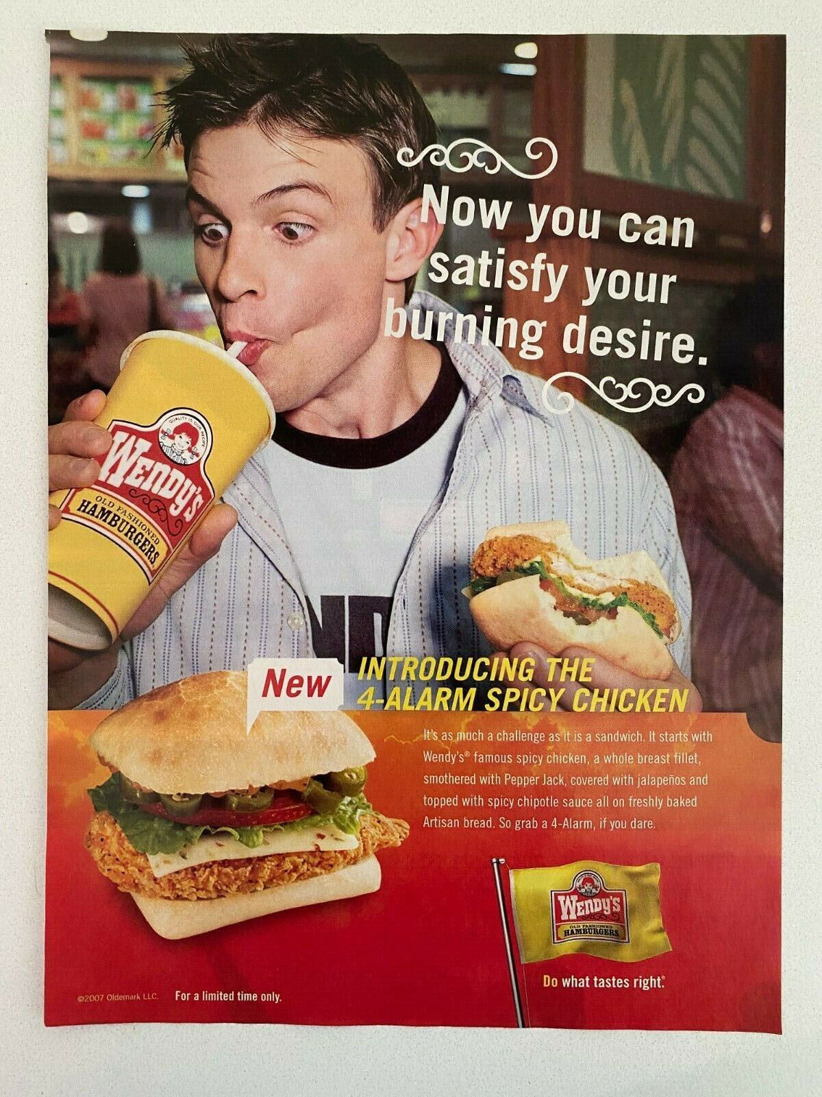 A young man drinks from a Wendys cup while holding a spicy chicken sandwich. The text reads, Now you can satisfy your burning desire and Introducing the 4-Alarm Spicy Chicken. Another sandwich lies on a table with a Wendys sauce packet.