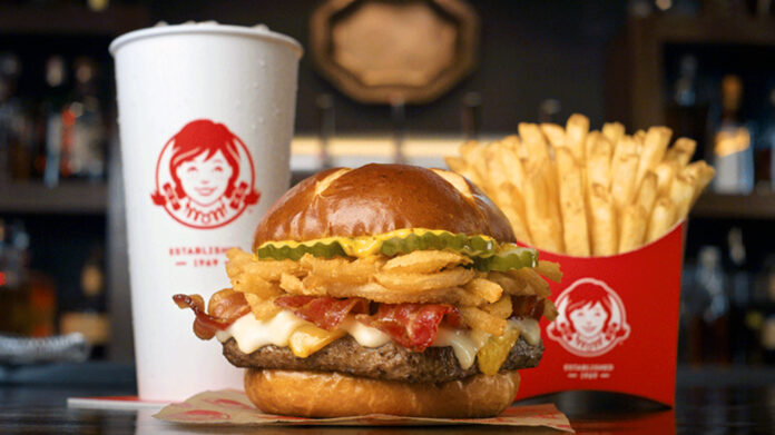 A burger with bacon, fried onions, pickles, and cheese in a pretzel bun is in front of a container of fries and a drink cup. The background is blurred, showing a restaurant setting.